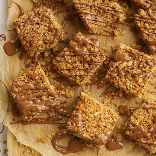 Squares of homemade flapjacks drizzled with chocolate on a wooden board and baking paper.