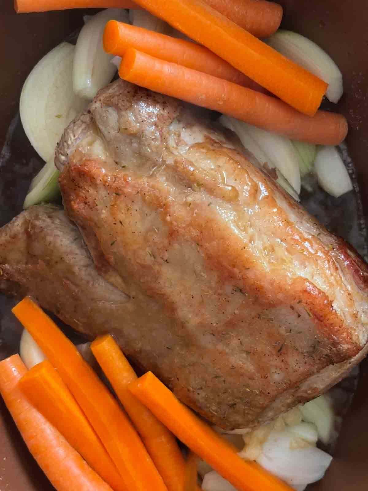 A browned beef joint next to carrots and onion, put in a slow cooker ready to be slow cooked.