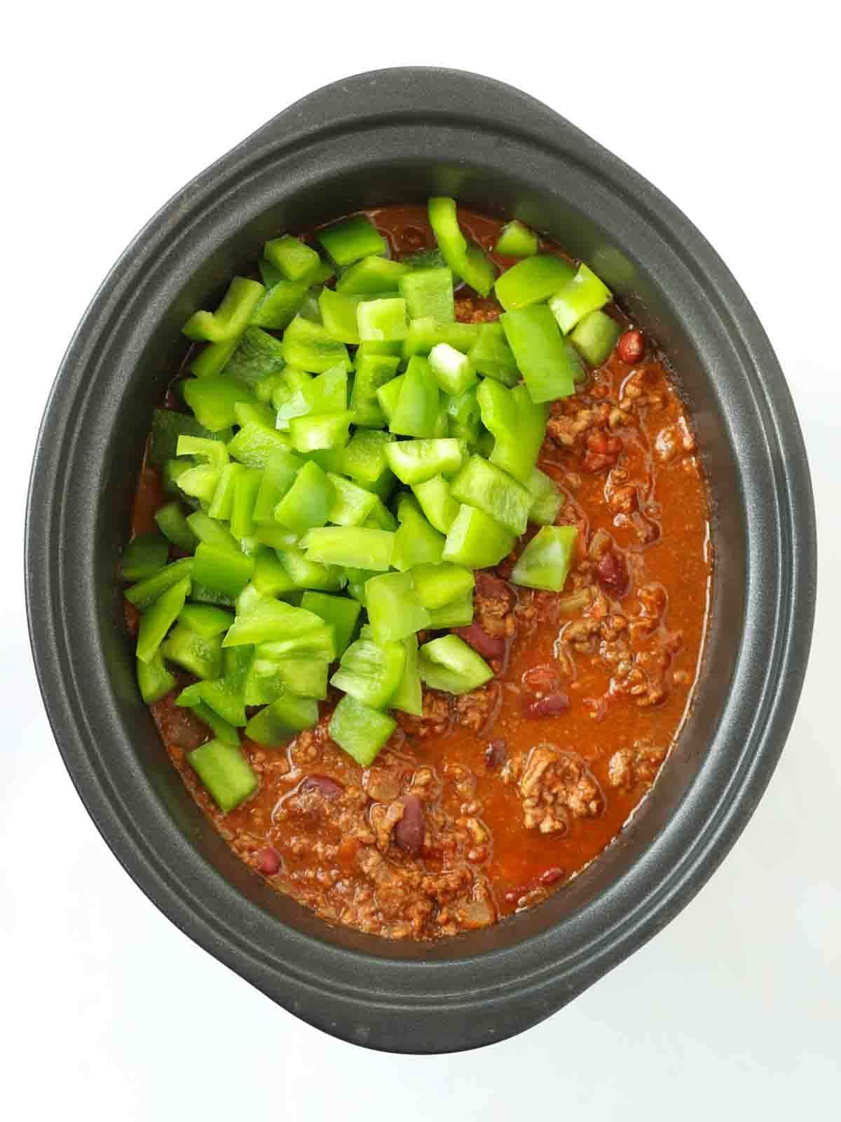 A slow cooker pan filled with slow cooker chilli, with raw green peppers on top, ready to go in.