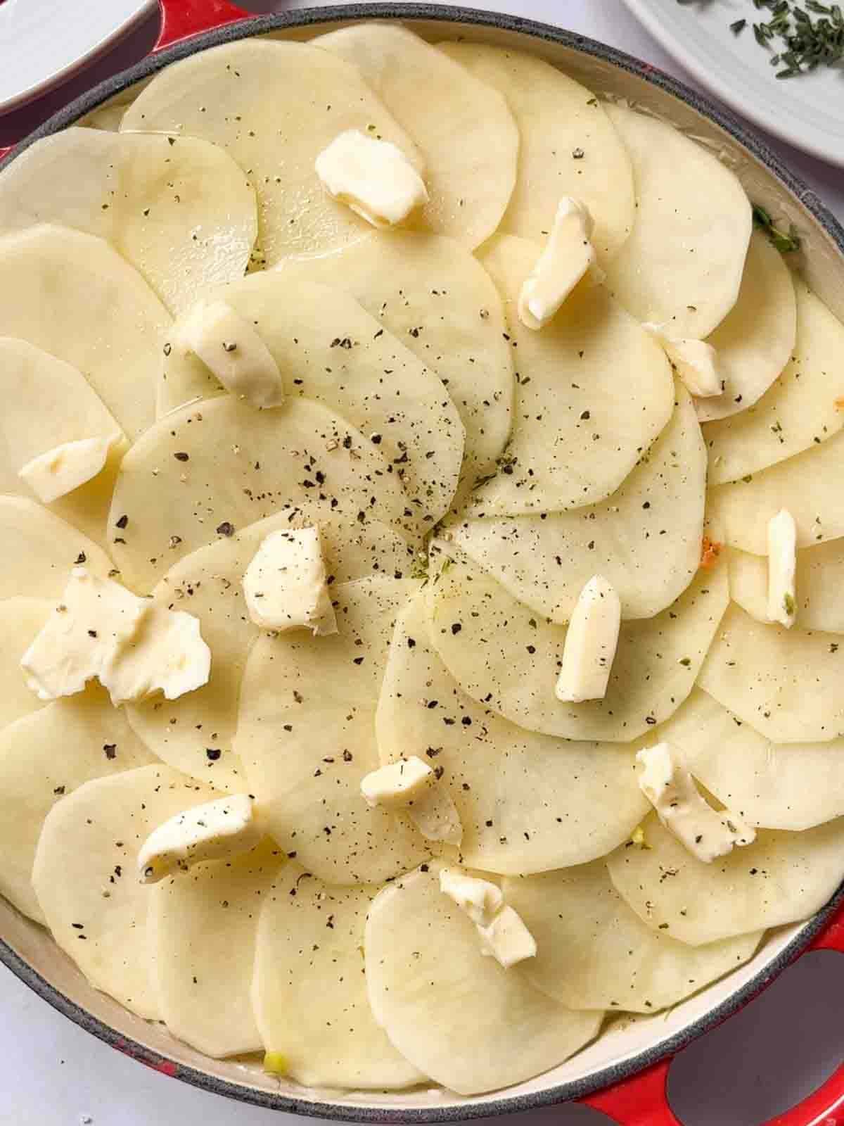 Rounds of sliced potatoes with knobs of butter and black pepper on top, for step 4 in the recipe for Boulangere Potatoes.