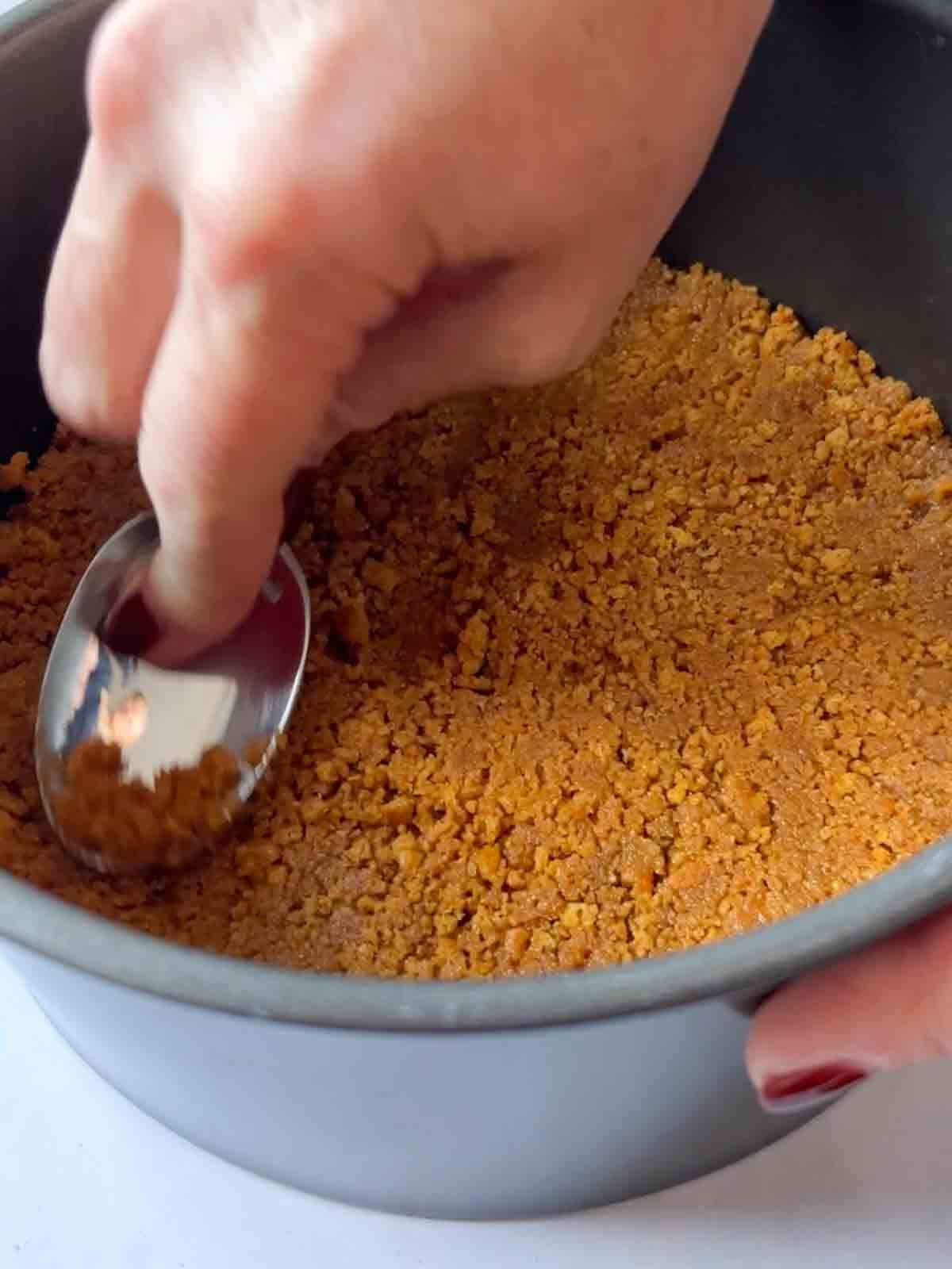 Crushed biscuits being pressed down into a tin with the back of a spoon for the recipe White Chocolate Cheesecake.