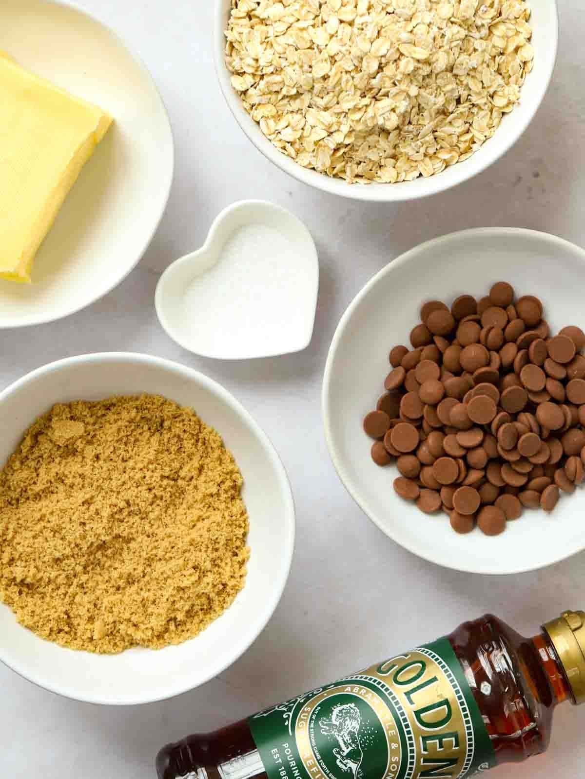 The raw ingredients for a flapjack recipe laid out on a counter, including a bottle of Golden Syrup, and bowls filled with oats, butter, sugar, salt and choc chips.
