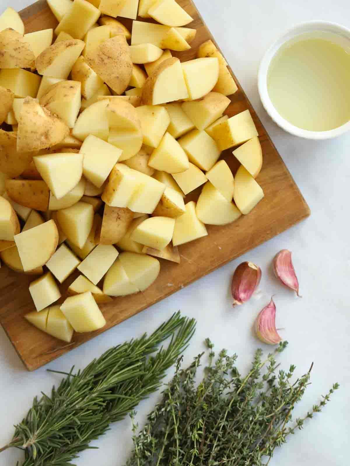 The raw ingredients for making parmentier potatoes laid out on a counter, including cubes of white potatoes, oil, garlic cloves and fresh herbs.