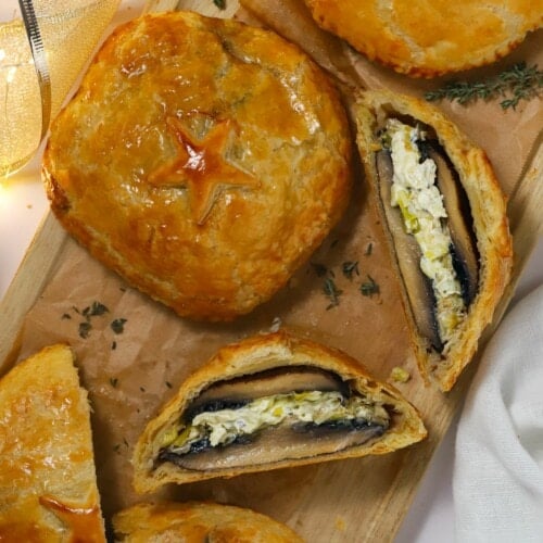 Four puff pastry mushroom wellingtons on a long copping board, with one sliced and on its side to show the filling inside.