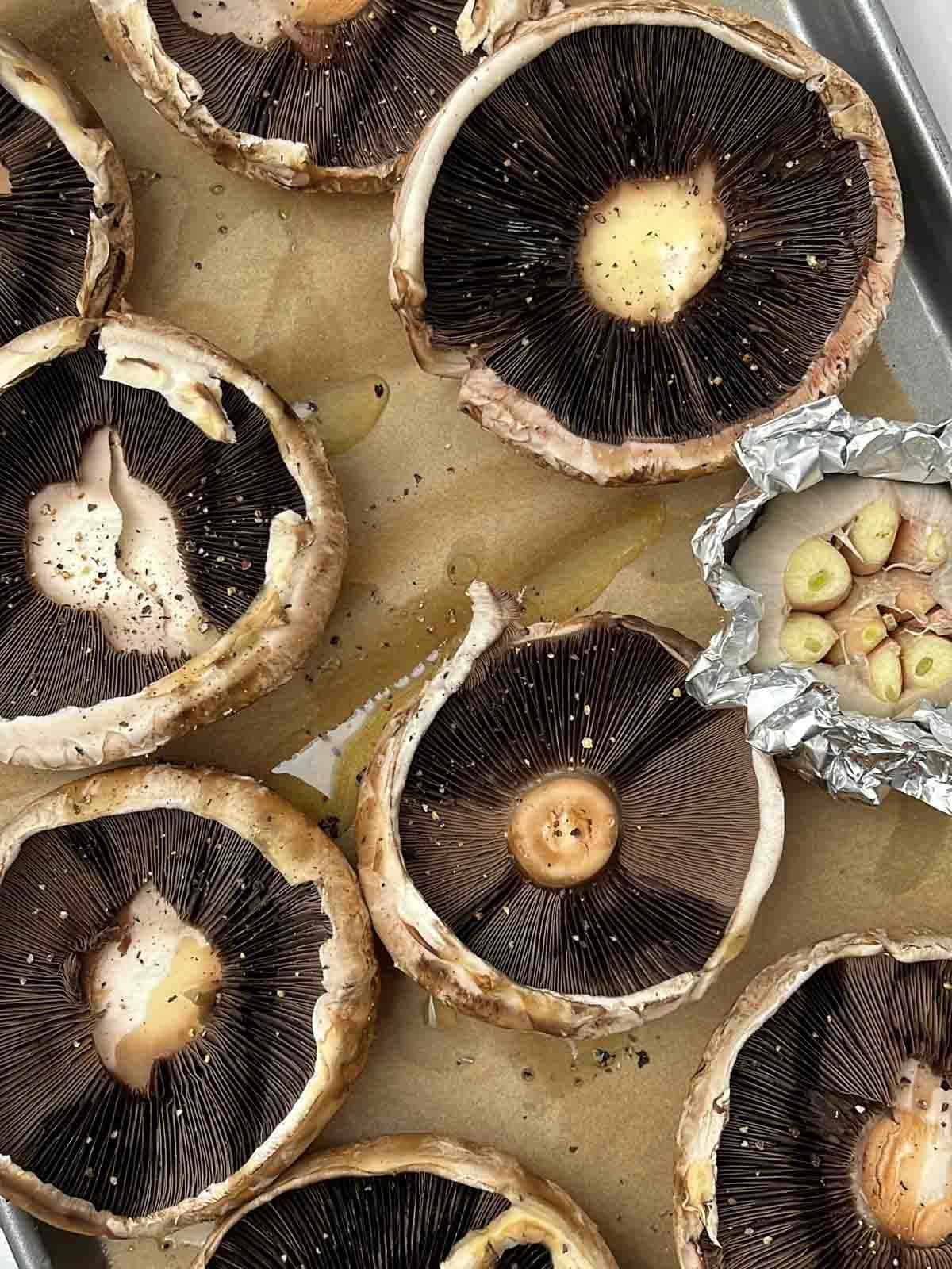 Portobello mushrooms and a garlic bulb drizzled with oil on a baking tray, ready to make mushroom wellingtons.