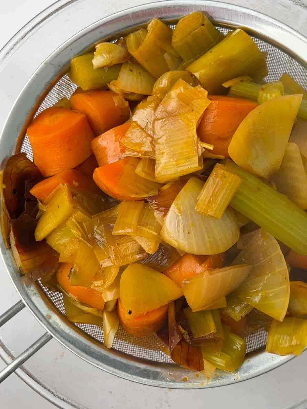 Cooked vegetables in a strainer over a bowl for step 3 in the recipe for vegetarian gravy.