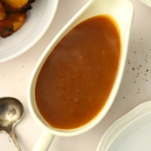 A gravy boat with vegetarian gravy inside, sitting on a white table with a spoon and potatoes just out of shot.
