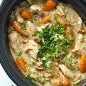 A chicken casserole in a slow cooker, ready to be served. With carrots, chicken, bacon bits in a creamy sauce, garnished with parsley.
