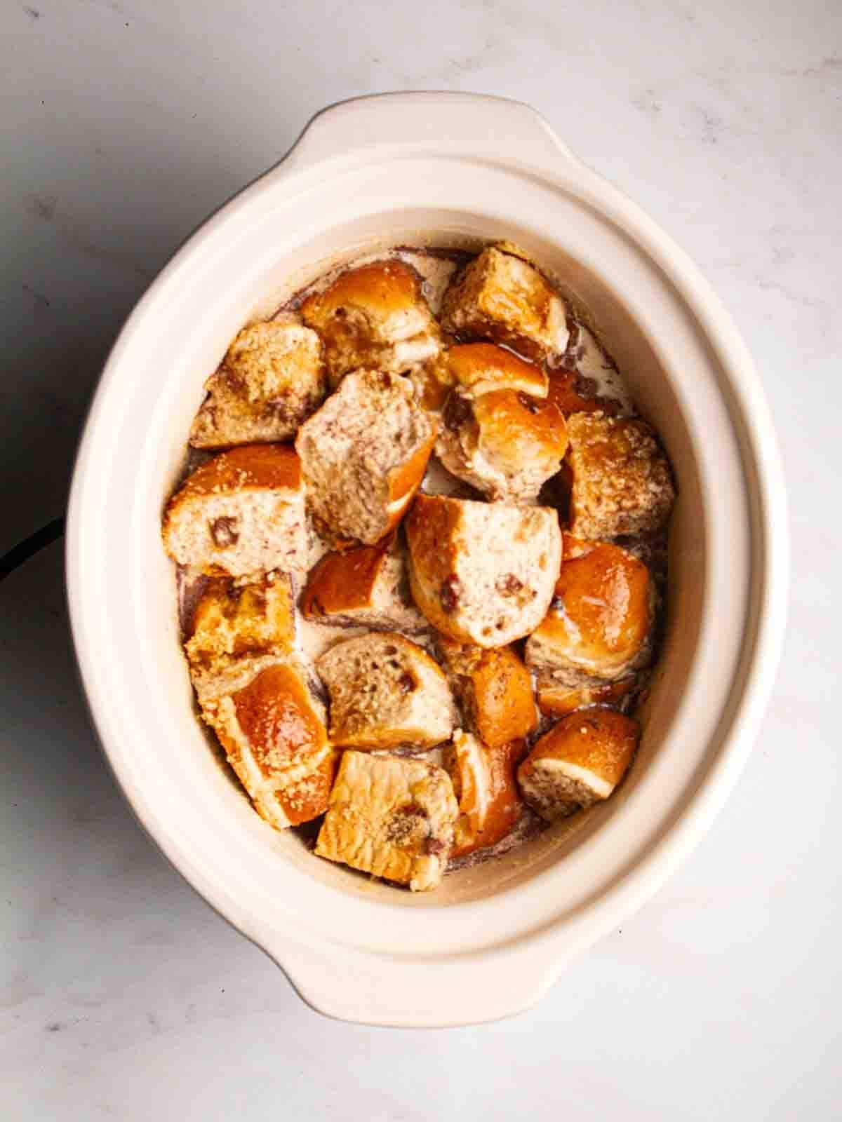A slow cooker pan filled with hot cross buns and liquid for step 2 in the recipe for Hot Cross Bun Bread and Butter Pudding.