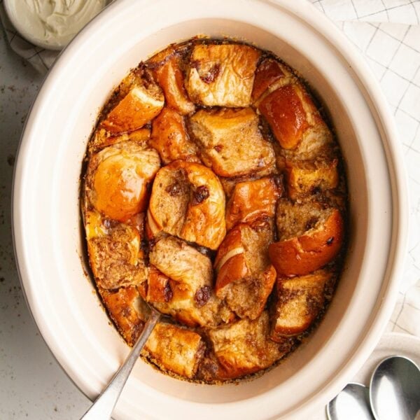 A big slow cooker pot filled with cooked bread and butter pudding with hot cross buns and a spoon sticking out of it.