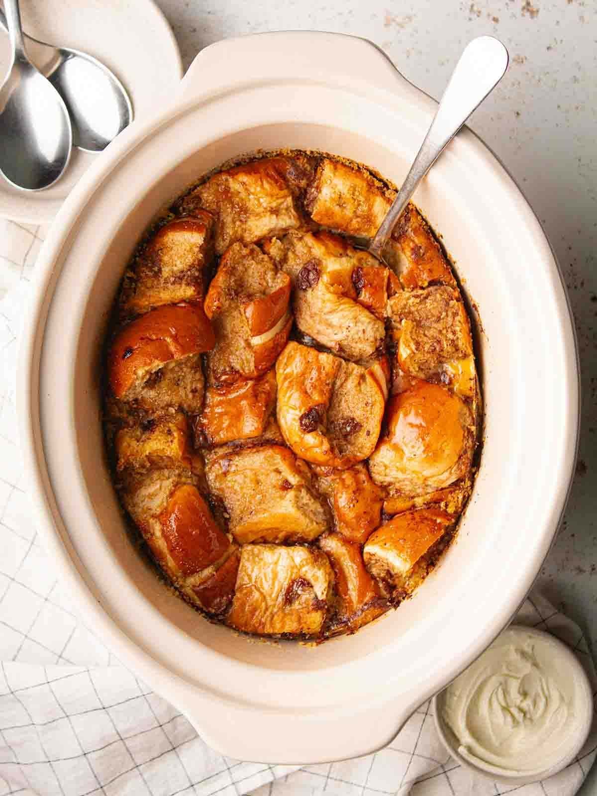 A slow cooker pan with a spoon inside, ready to dish up the Hot Cross Bun Bread and Butter Pudding inside.