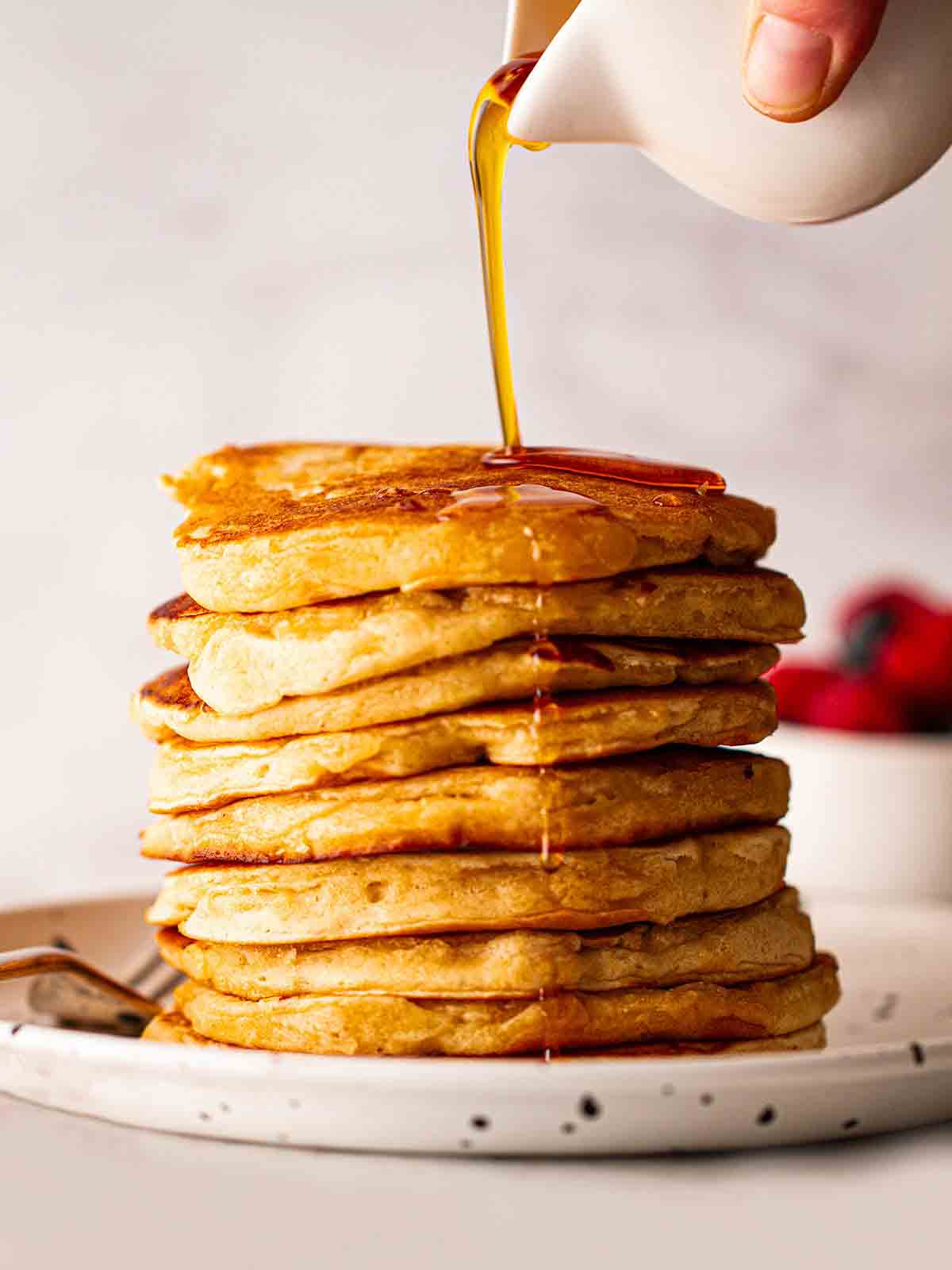 A big stack of American Pancakes on a plate with maple syrup being drizzled over the top from a jug.
