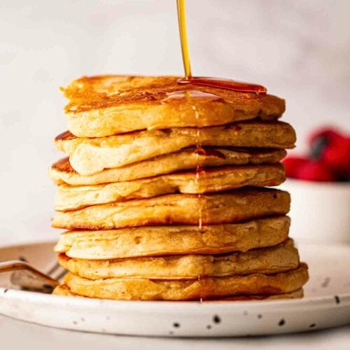 Maple syrup being drizzled over a big pile of golden American Pancakes.