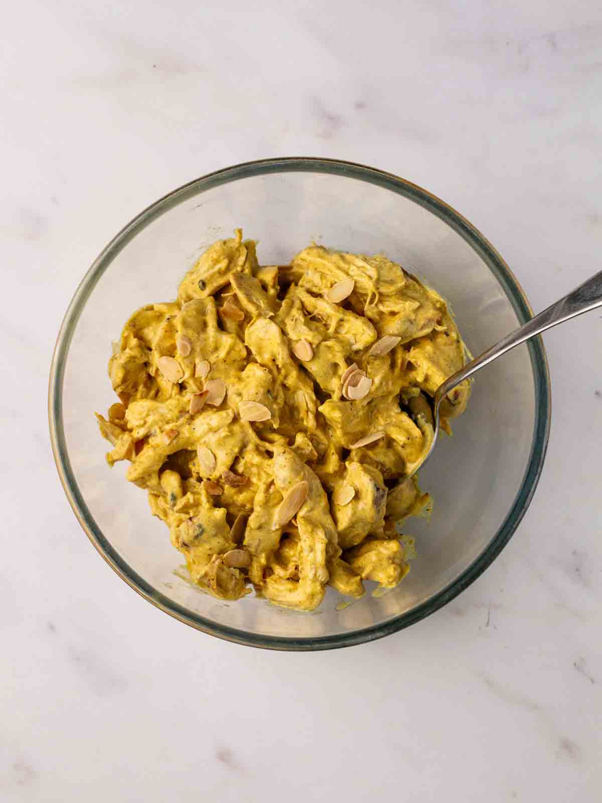 A glass bowl filled with Coronation Chicken chunks and flaked almond, with a spoon inside, stirring.