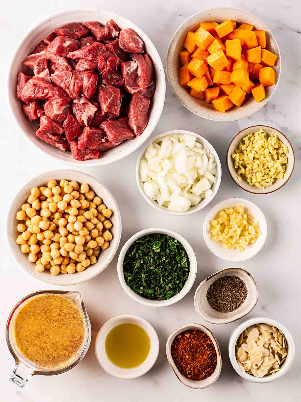The raw ingredients for a lamb tagine laid out in bowls on a white counter top.