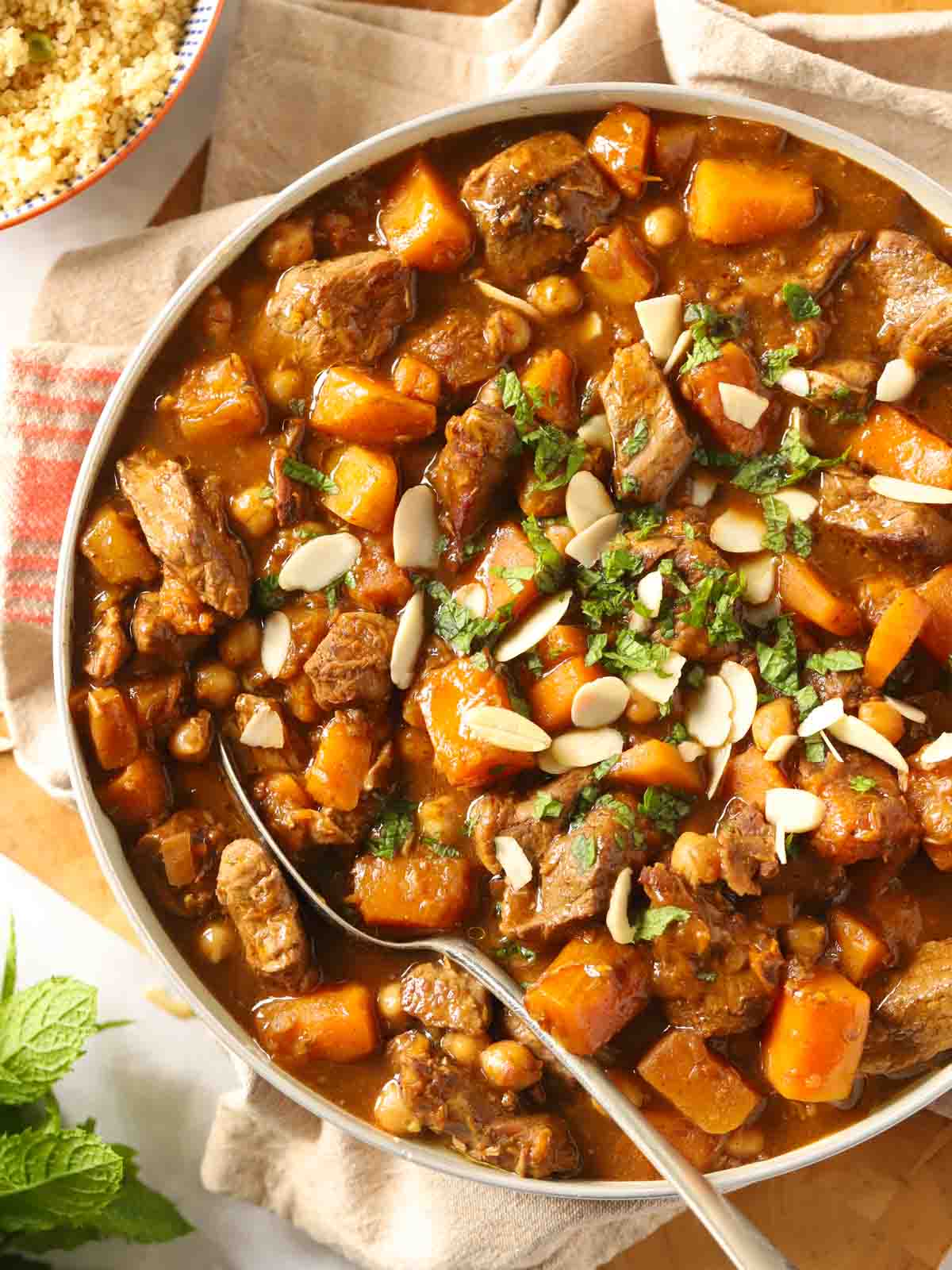 A bowl of lamb tagine with chunks of lamb and butternut squash, on a table with a tea towel, couscous and a silver spoon ready to dish it up.