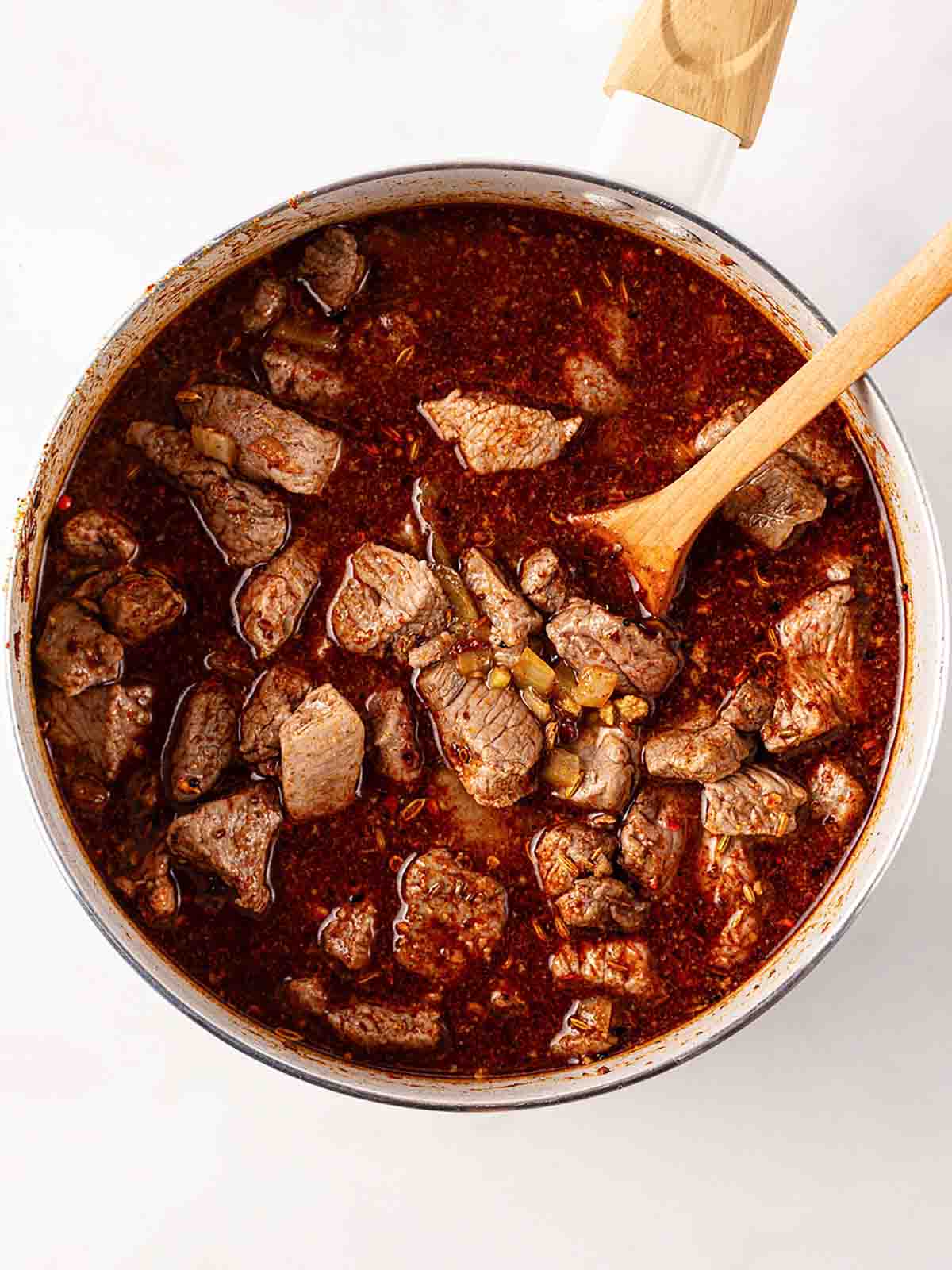 A big pan filled with lamb chunks in a red watery sauce with a wooden spoon, for step 3 in the recipe for Lamb Tagine.