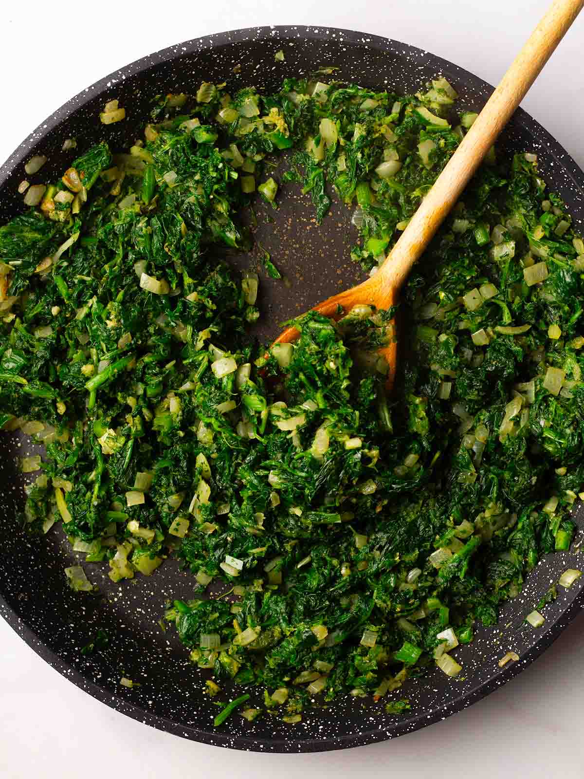 A frying pan filled with bright green spinach and onions with a wooden spoon stirring, for step 3 in the recipe for Palak Paneer.