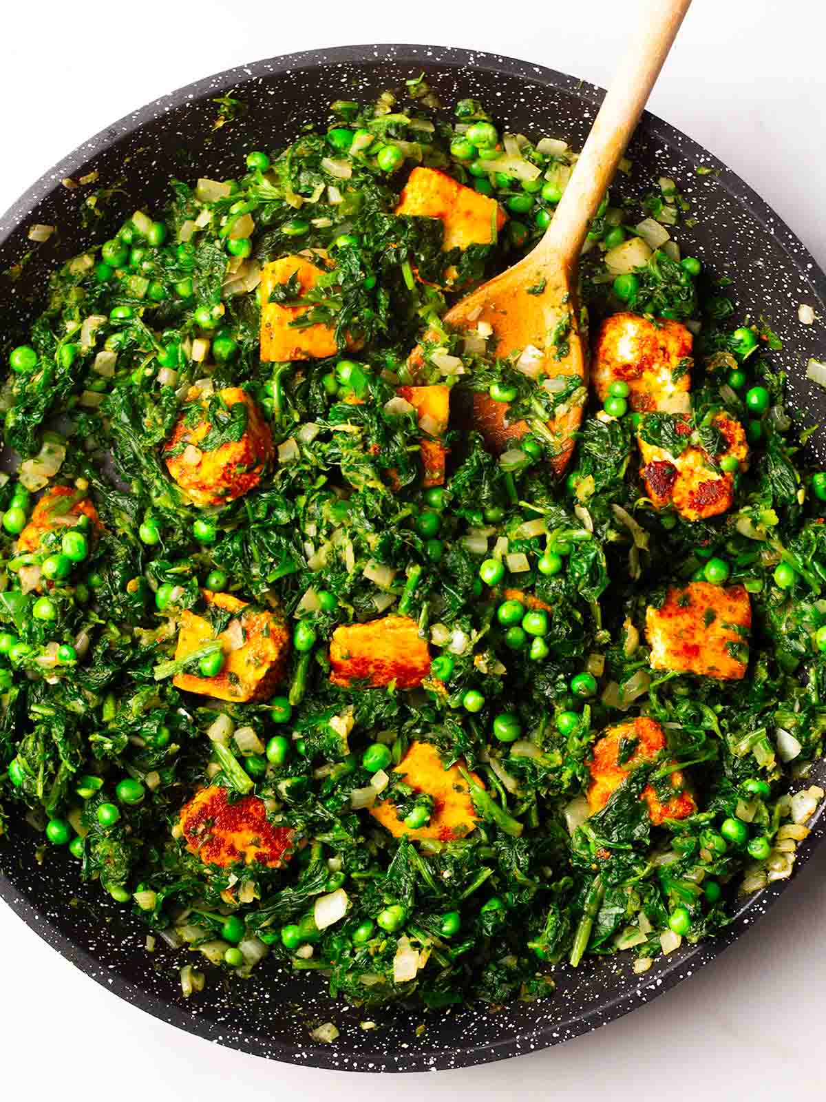 A frying pan with a combination of ingredients being stirred by a wooden spoon for step 4 in the recipe for palak paneer.