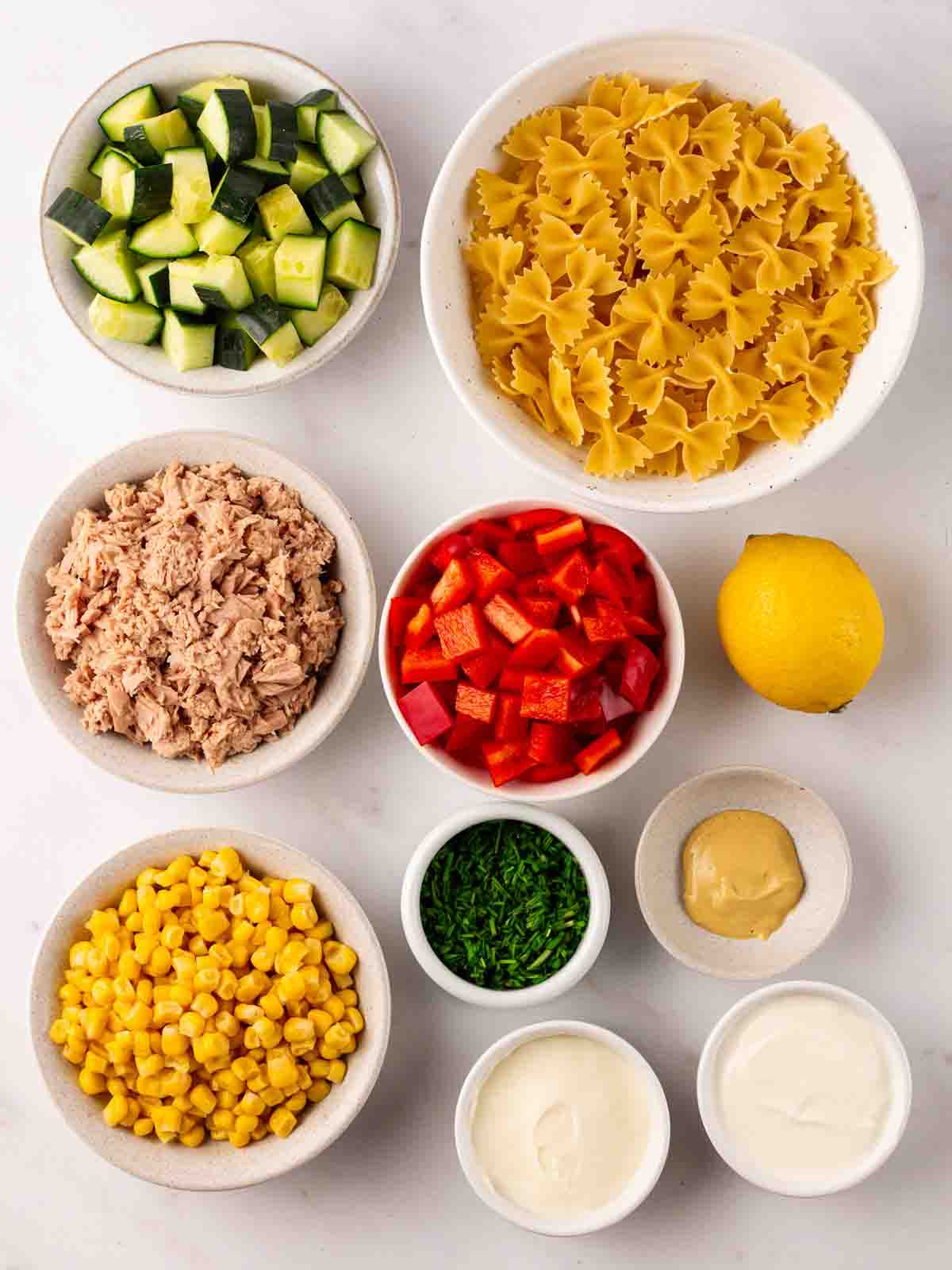 Ingredients for Tuna Pasta Salad with pasta, cucumber, peppers and corn in little bowls on a counter top.