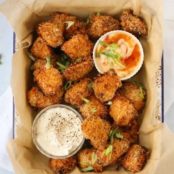 Coated golden homemade chicken nuggets in a baking dish with two pots of dips at the side.