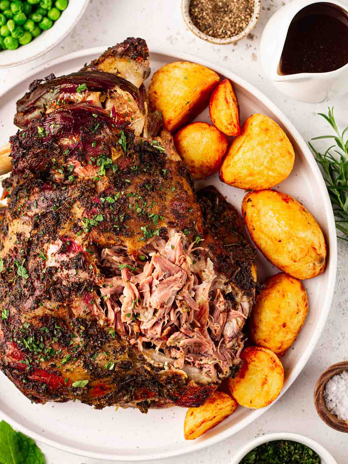 A shoulder of roasted lamb with a green herby crust, on a white plate with roast potatoes around the edge and peas and gravy off to the side.