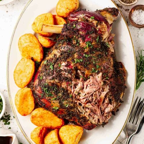 A white serving dish with a lamb roasted shoulder, next to roast potatoes on a table.