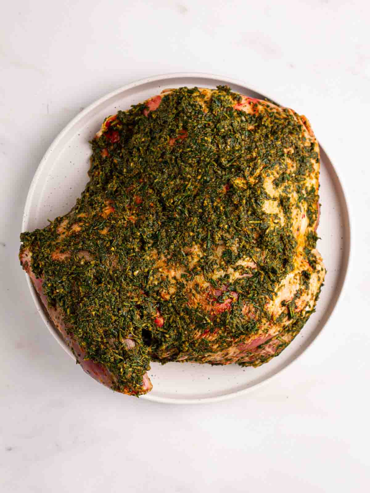 A raw shoulder of lamb with a green herby crust on top on a plate, ready to go in the oven for the recipe Slow Roasted Lamb Shoulder.