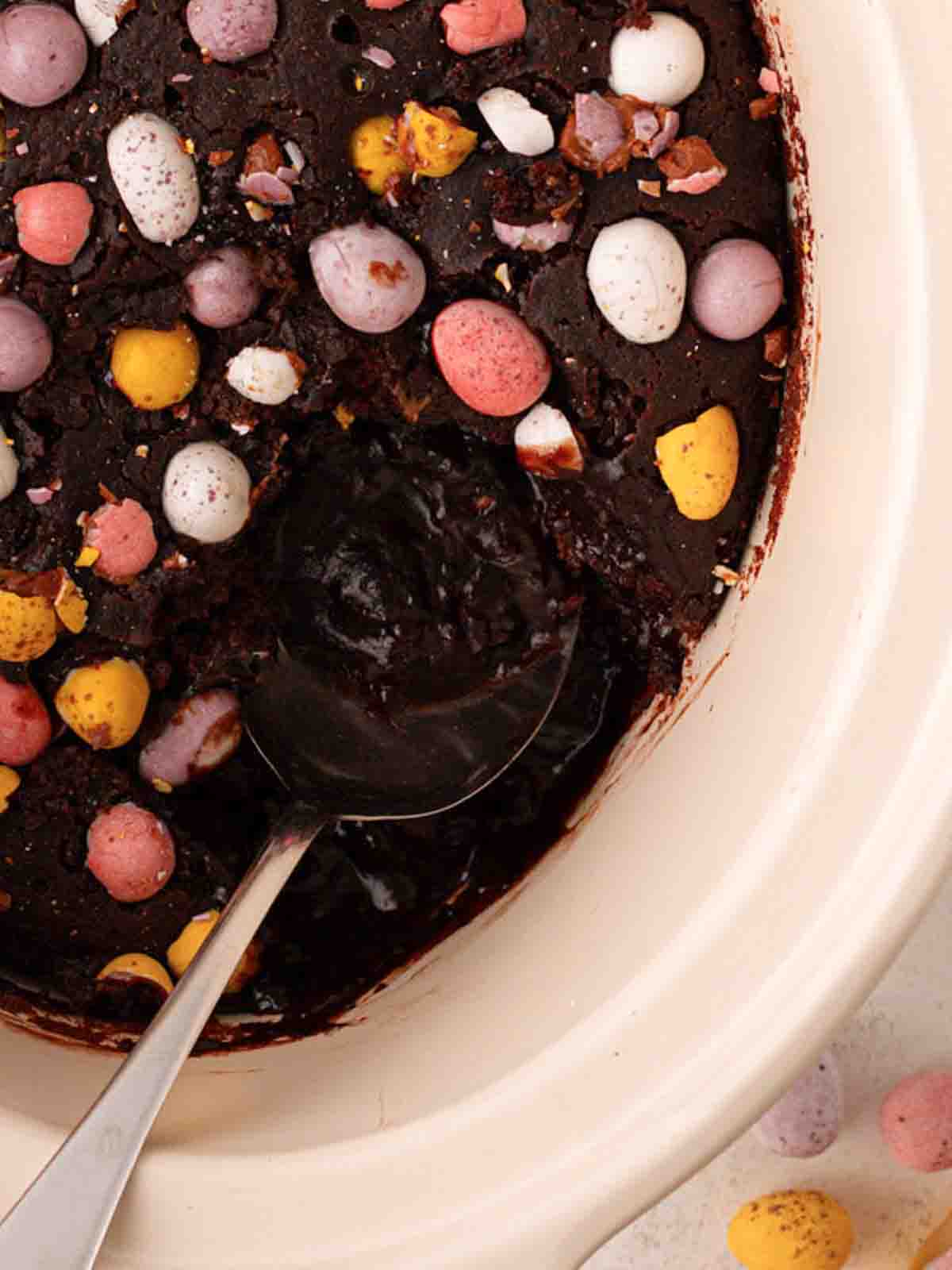 A spoon in a chocolate slow cooker pudding, revealing a gooey inside and mini eggs on top.