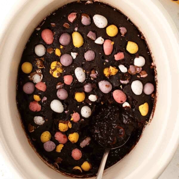 A slow cooker filled with cooked chocolate easter pudding with mini eggs on top and a spoon about to serve up.