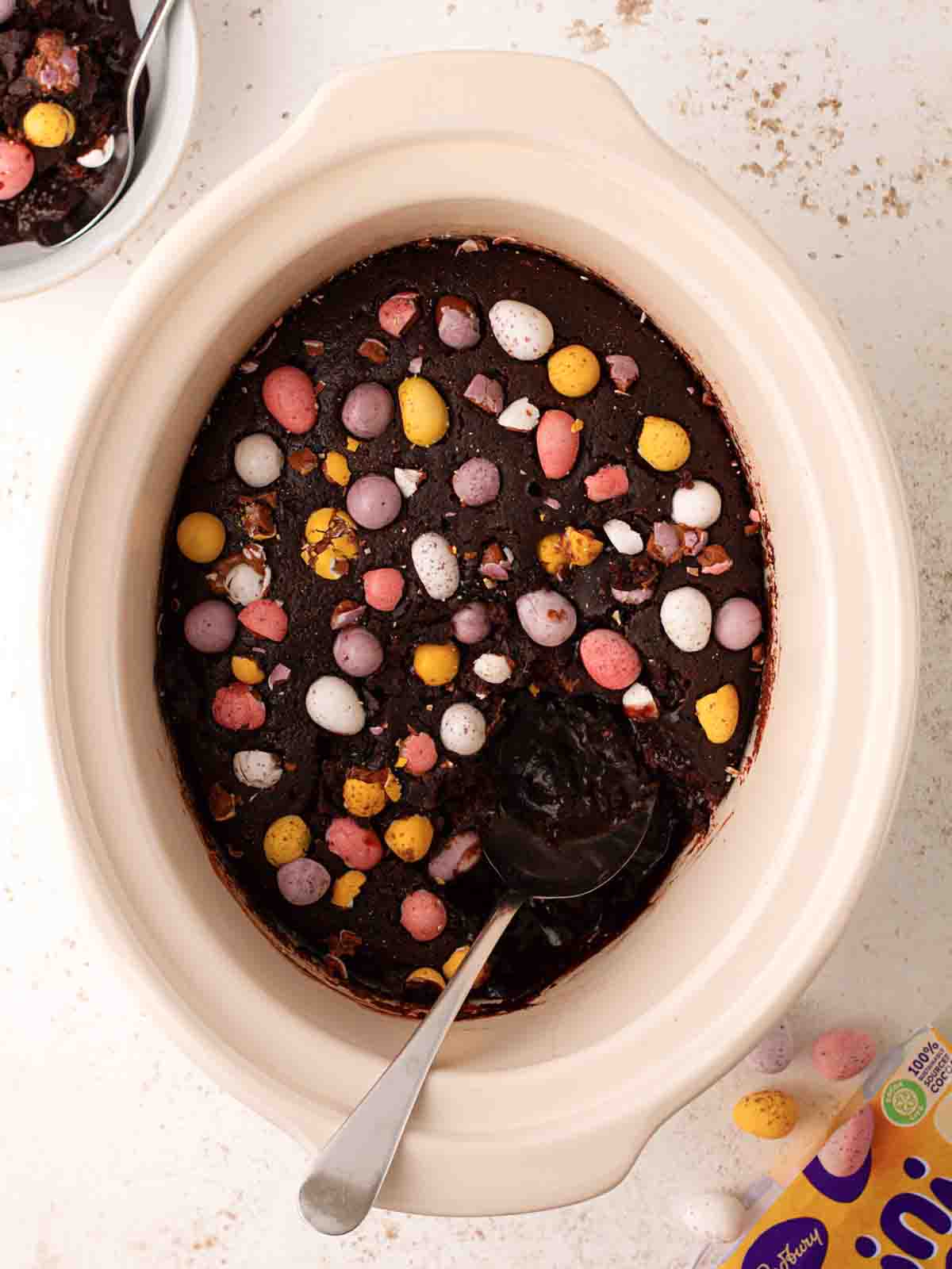 Looking down on a table with a slow cooker pan filled with mini egg easter chocolate pudding with a bowl at the side and a bag of mini eggs.