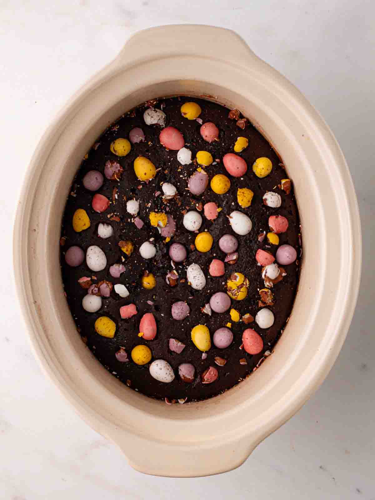 A slow cooker pan with mini eggs set into a chocolate pudding for step 6 in the recipe for Slow Cooker Mini Egg Easter Pudding.