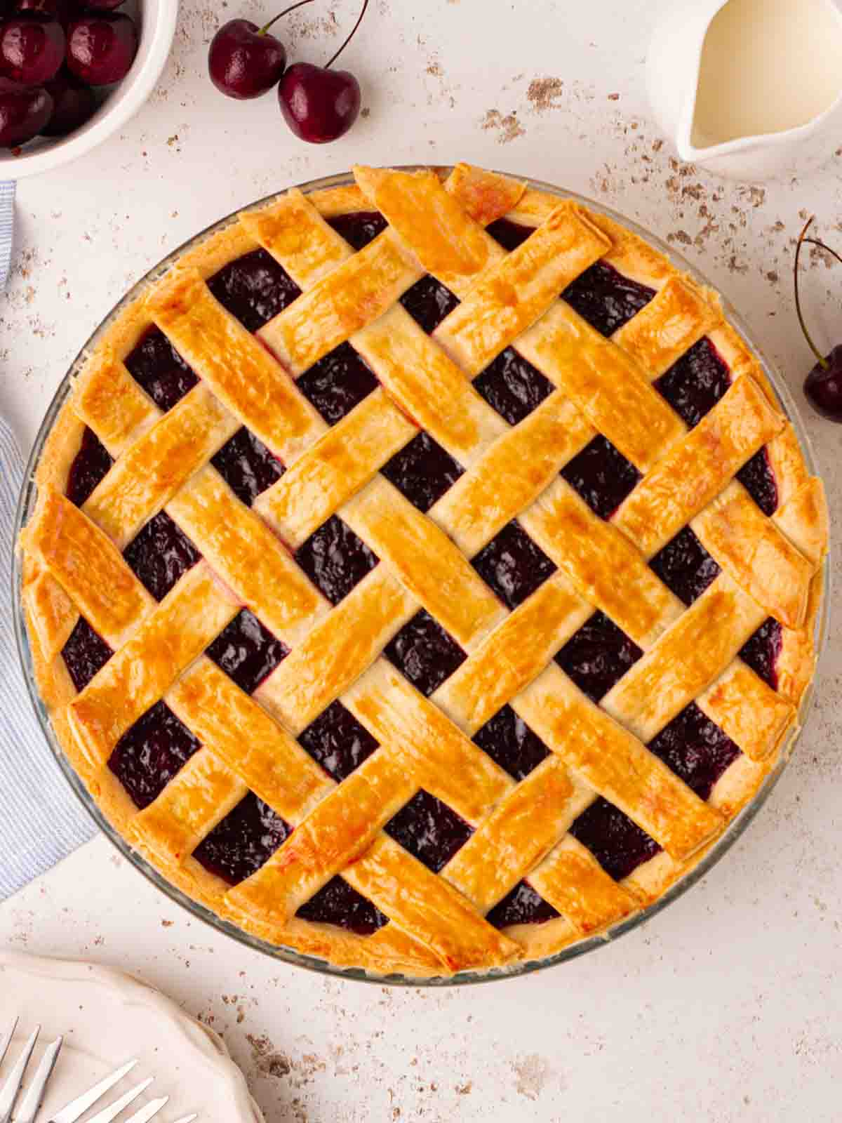 A full cherry pie on a table with a golden topping, and cream and cherries to the side.