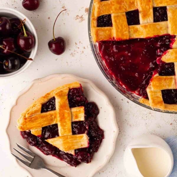 A homemade cherry pie with a lattice topping, with a portion on a plate, ready to eat.