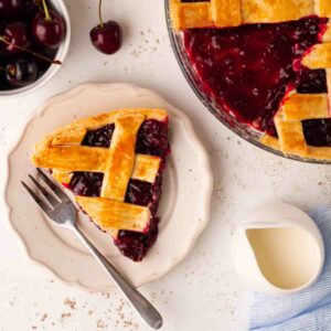 A slice of cherry pie on a plate with a fork, with the rest of the pie to the side, and a bowl of cherries, and a jug of cream on the side.