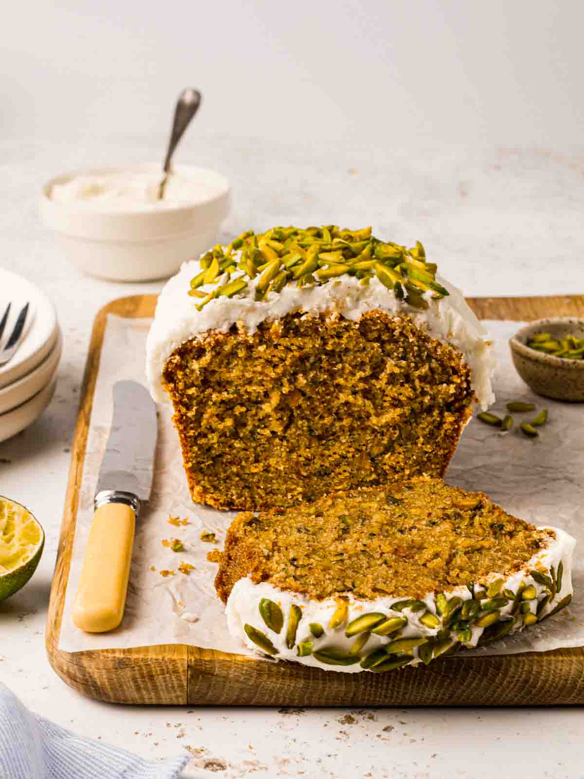 A sliced Courgette Cake from the side, on a board with a knife to the side and bowls and cutlery too.