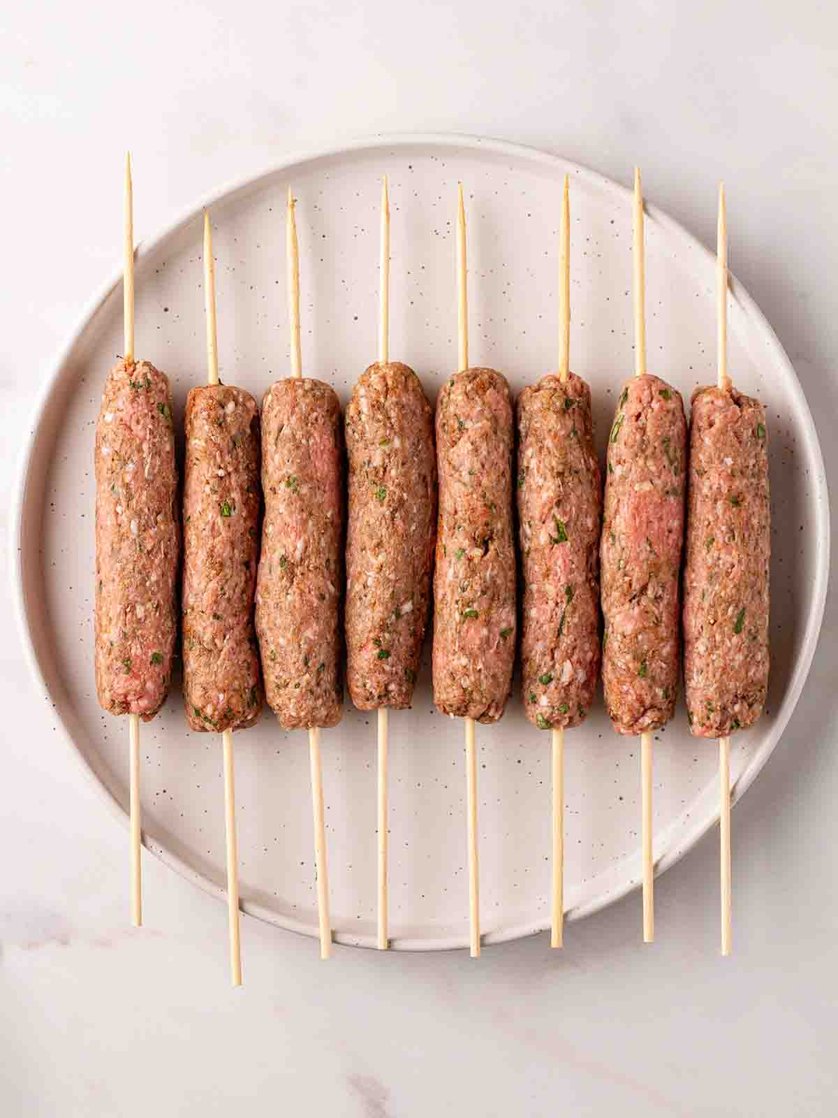 Raw lamb kofta kebabs with skewers through the middle, on a plate, ready to be cooked.