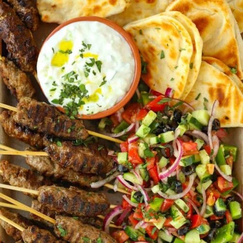A tray with lamb kofta kebabs on with skewers, served with tzatziki dip in the middle and flatbreads.