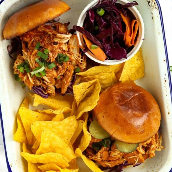 A dish with two filled buns with slow cooker pulled chicken in, ready to eat, with tortilla chips on the side and coleslaw.
