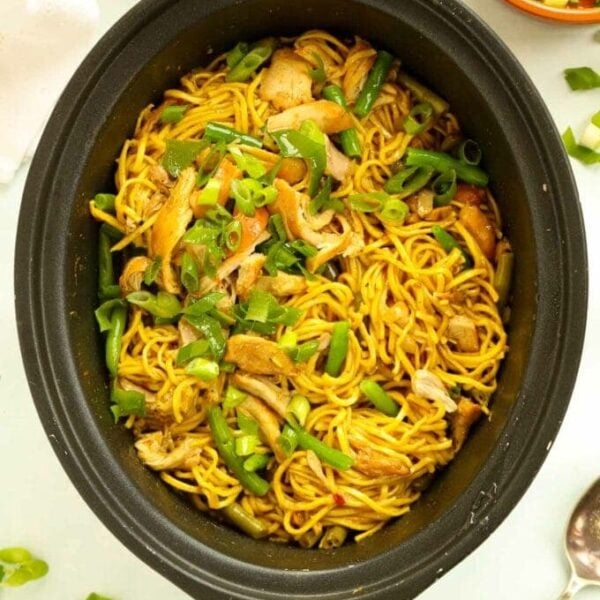 A black slow cooker pan filled with curried noodles and chicken, with green beans.