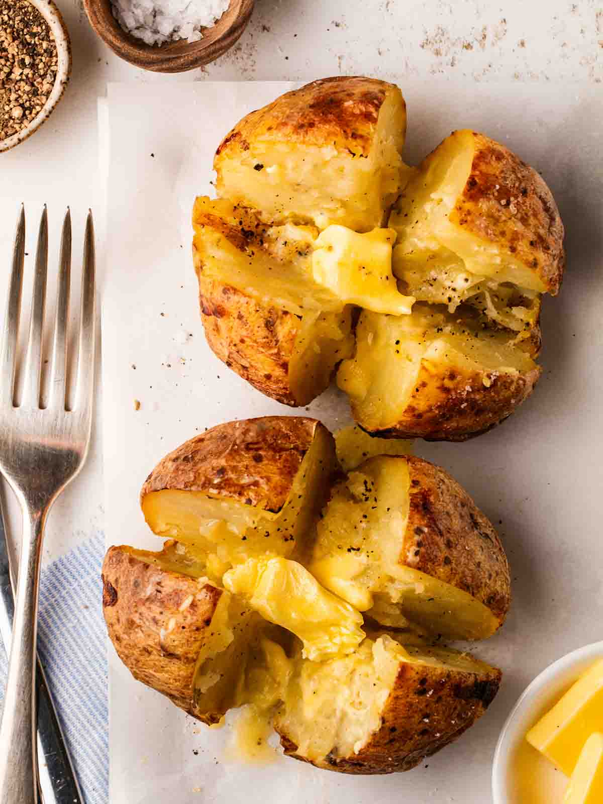 Two jacket potatoes having been cooked in the air fryer with butter melting in the centre, cut into four with a fork at the side.