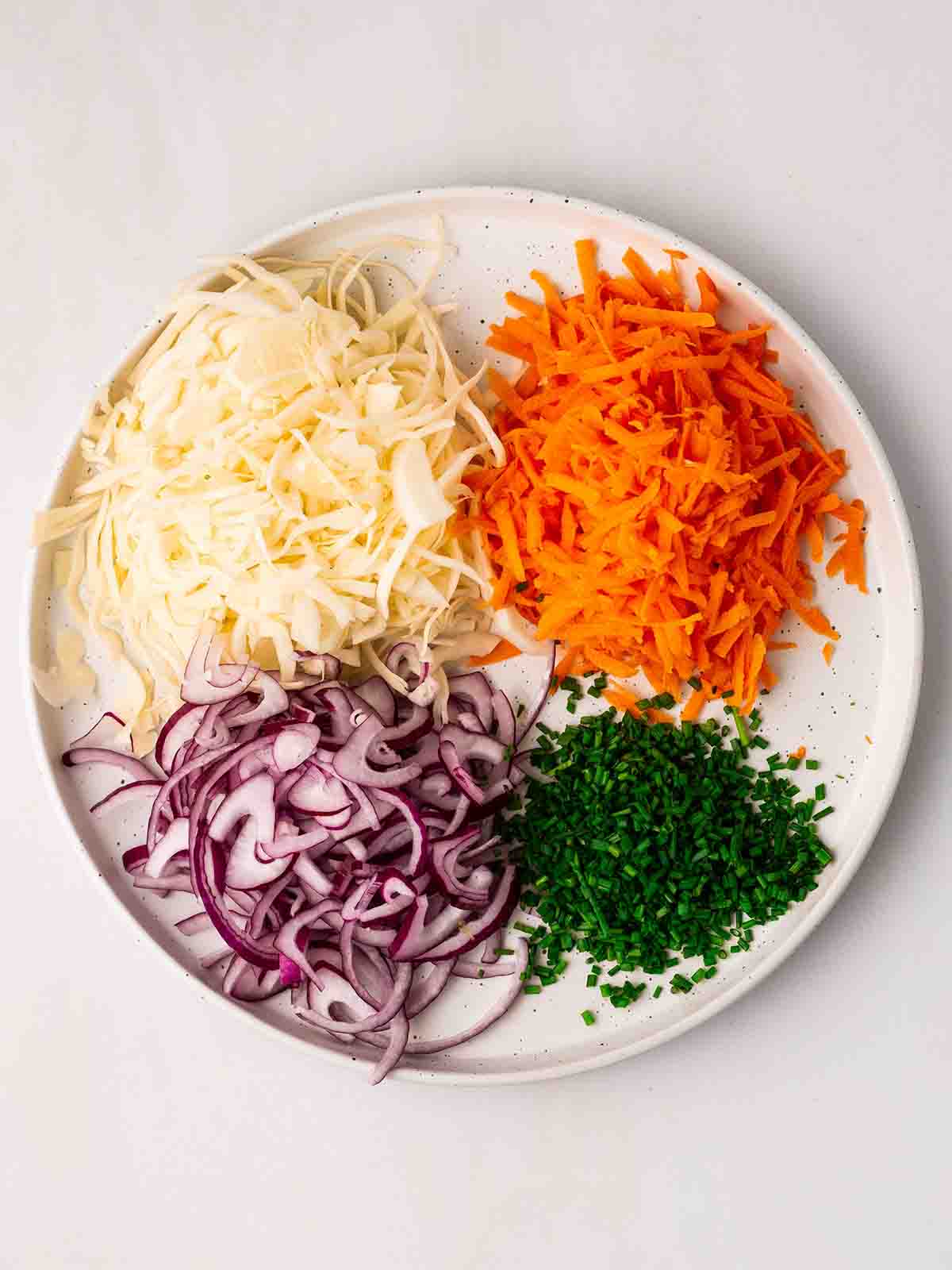 A plate filled with white cabbage, grated carrot, finely sliced red onion and chopped chives, for step 2 in the recipe for homemade coleslaw.