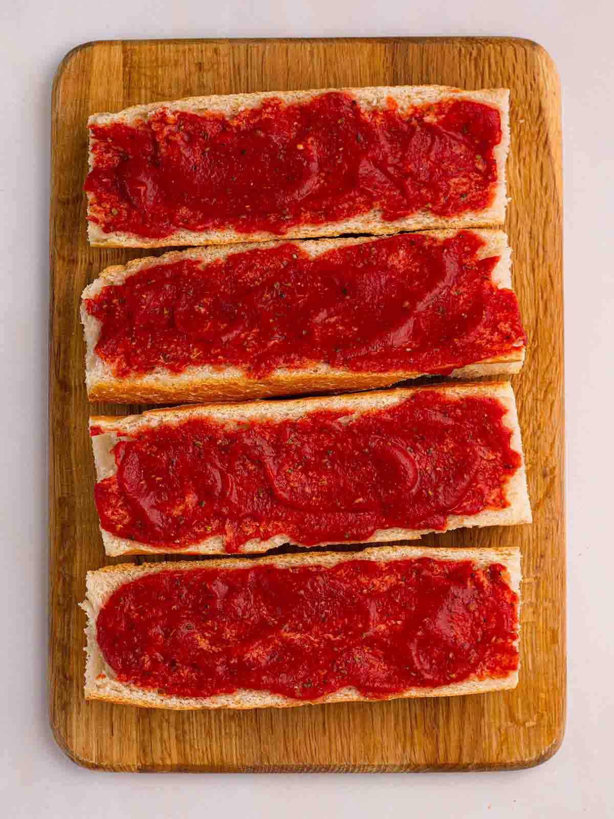 Tomato passata spread on top of halved baguettes on a chopping board for step 3 in the recipe for French Bread Pizza.