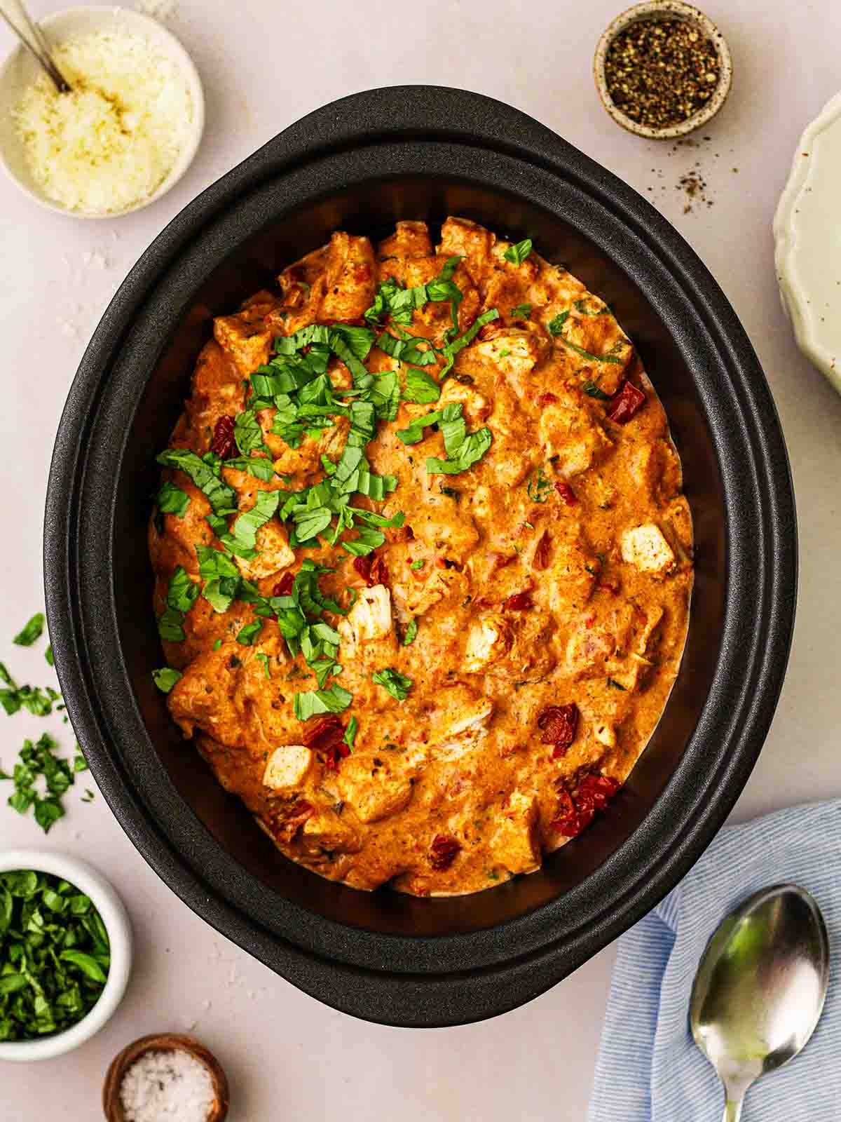 A black slow cooker pan filled with cooked Slow Cooker Marry Me Chicken, with small condiments around it and spoons.