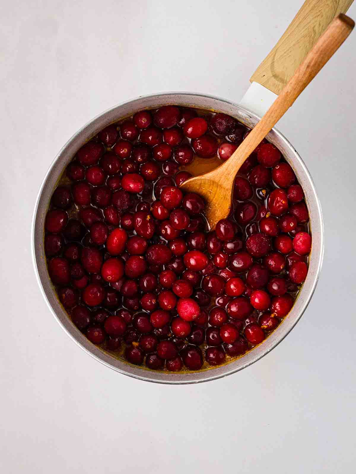 A pan with a wooden spoon stirring cranberries for step 1 in the recipe for Cranberry Sauce.