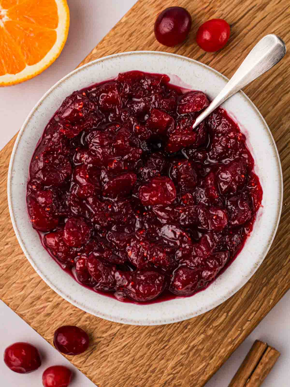 A board with a white bowl filled with cranberry sauce, with cranberries and an orange at the side.