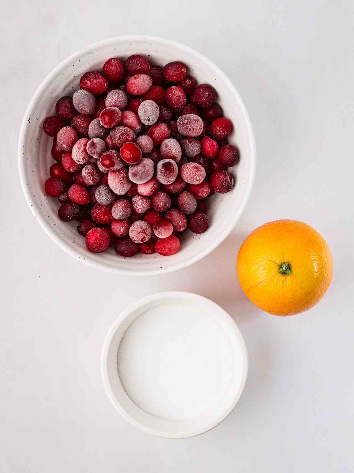 The ingredients for making cranberry sauce laid out on a counter top, including a bowl of frozen cranberries, an orange and sugar.
