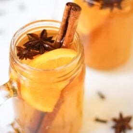 A close up of a glass jar of Mulled Cider, garnished with cinnamon stick, star anise and a slice of orange.