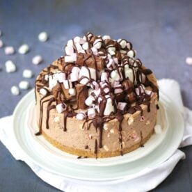 A Rocky Road Cheesecake on a white plate, piled high with marshmallow and chocolate bar topping and melted chocolate.