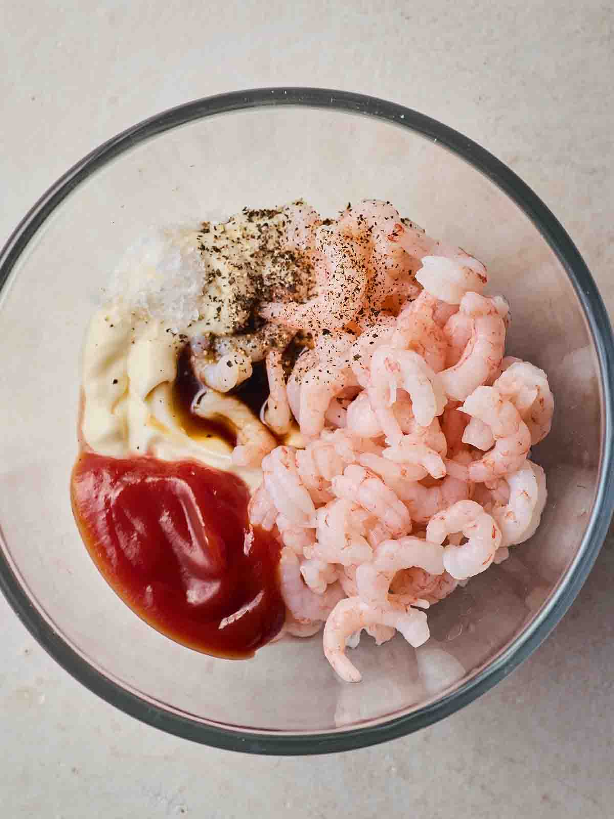 A glass bowl filled with cooked prawns, marie rose sauce ingredients and salt and pepper, ready to be mixed into a Prawn Cocktail filling.